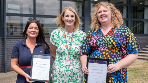 Three people standing in front of a building with large windows; two hold framed certificates or awards